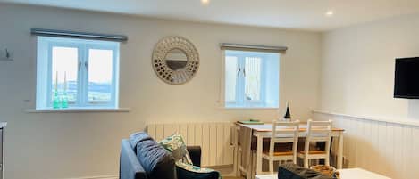 En stue med sofa, spisebord med stole, to vinduer med gardiner, et vægmonteret tv og en radiator.