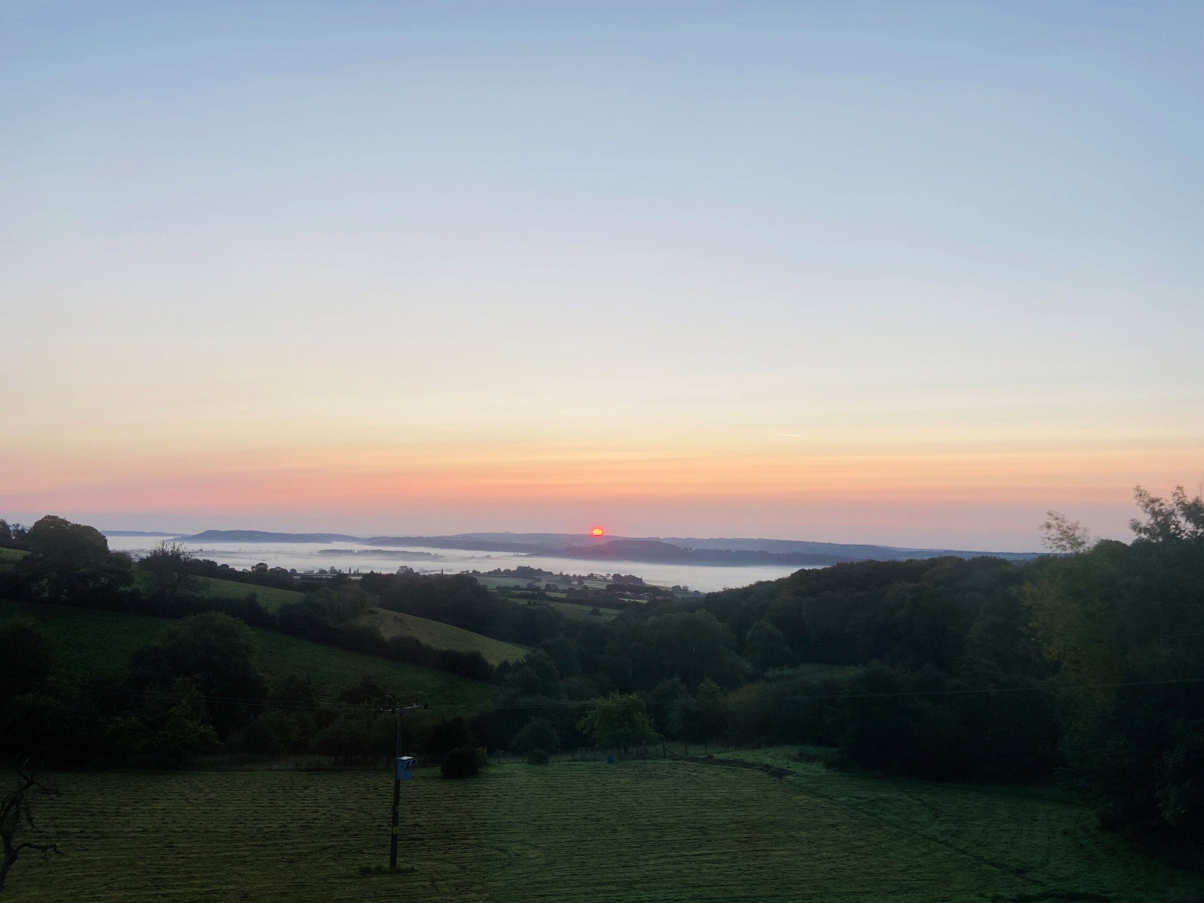 A rural landscape with rolling hills and a sunset over a body of water.