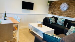 Uma sala de estar moderna com uma parede de destaque em pedra, uma televisão de tela plana, uma área de jantar com mesa e cadeiras de madeira, e um sofá preto com almofadas estampadas.