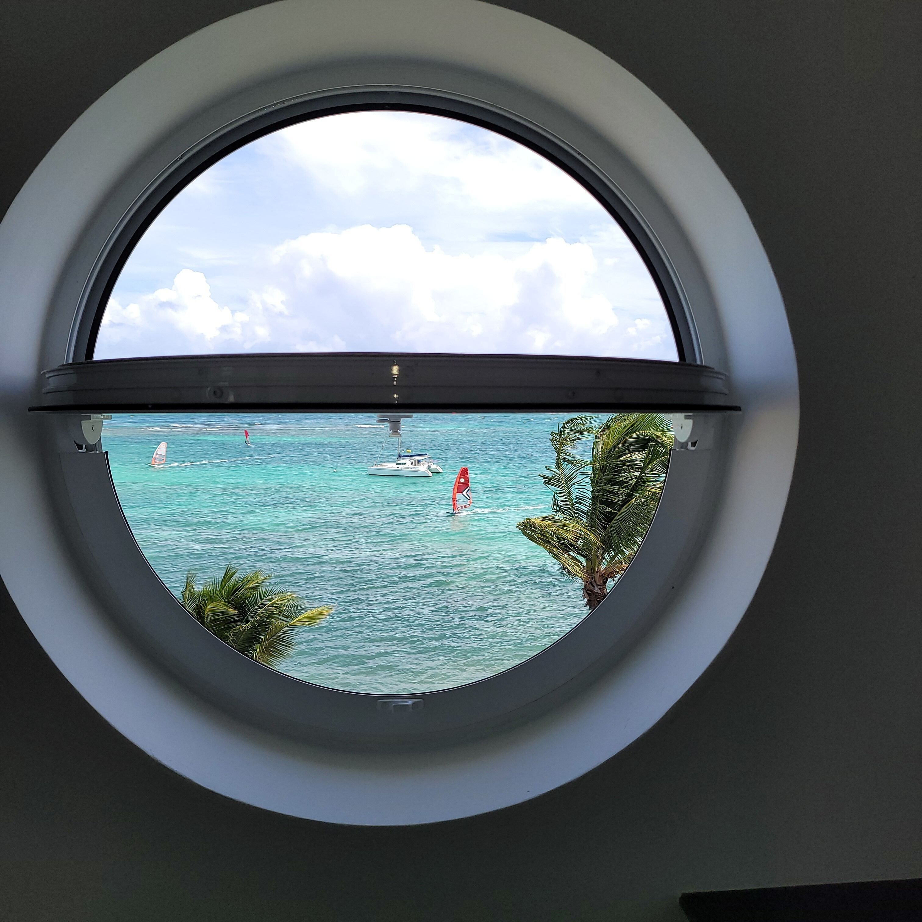 A round window with a view of turquoise water, boats, and palm trees.