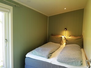 Quarto pequeno com cama de solteiro, janela, mesa de cabeceira e luminária na parede.