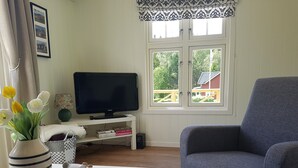 A living room with a television on a white stand, a grey armchair, a white table with a tray of decorative items, and a window with a view of a wooden deck and greenery.