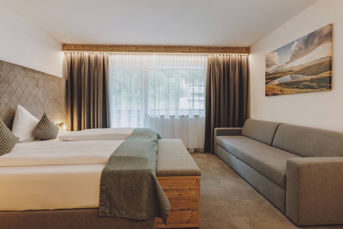 Quarto de hotel com cama, sofá, um banco de madeira e uma grande janela com cortinas.