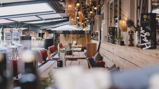 Un espacio al aire libre con mesas y bancos de madera, luces colgantes y vistas a la calle.