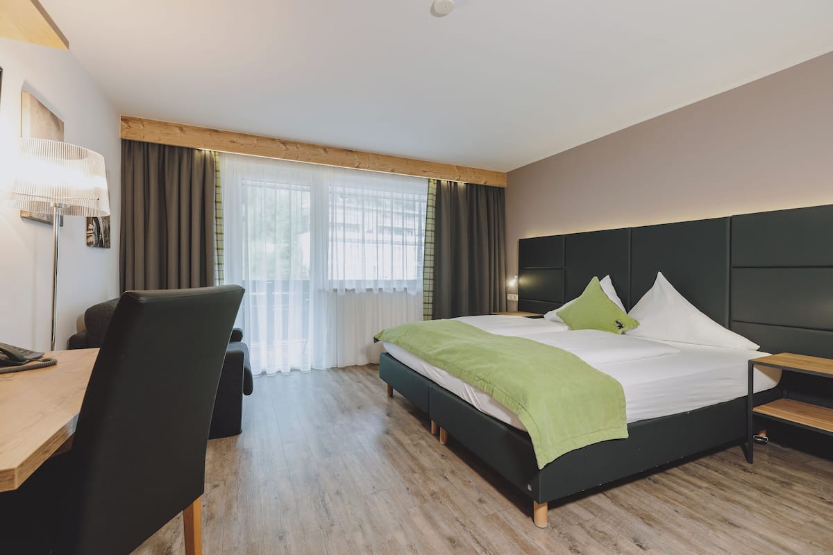 Quarto de hotel moderno com escrivaninha de madeira, uma cadeira confortável, uma cama grande com manta verde e cabeceira escura.