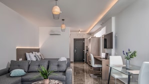 A modern living room with a grey sectional sofa, a glass coffee table, and a potted plant. There is a dining area with a glass table and white chairs. A TV is mounted on the wall, and a door leads to a hallway. - Ebenus Suites by Checkin (Hersonissos)