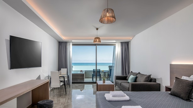 A modern hotel room with a large bed, a desk, a sofa, and a TV. There is a balcony with a view of the sea. - Ebenus Suites by Checkin (Hersonissos)