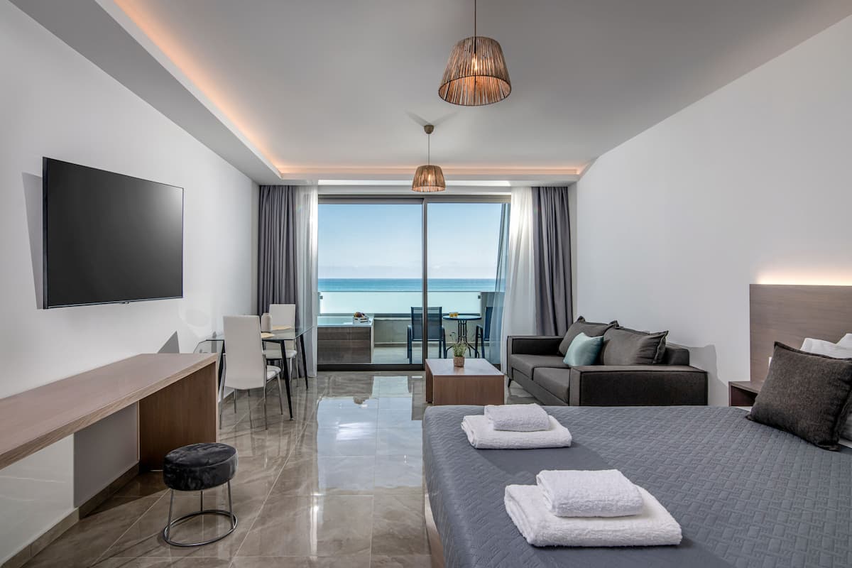 Una habitación de hotel moderna con una cama grande, un escritorio, un sofá y un televisor. Hay un balcón con vistas al mar.