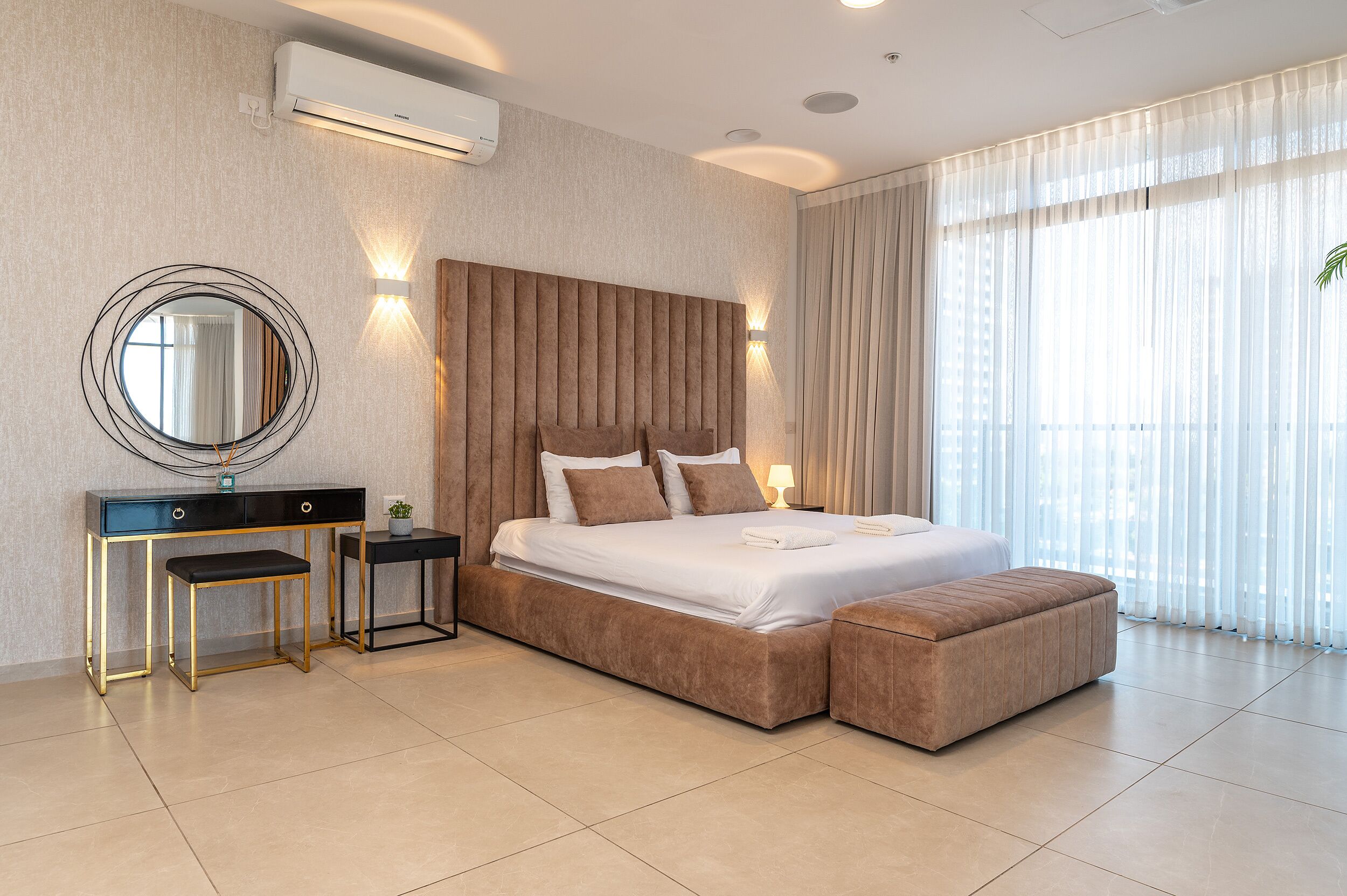 A modern bedroom with a large bed, bedside tables, a desk, and a large window with curtains.