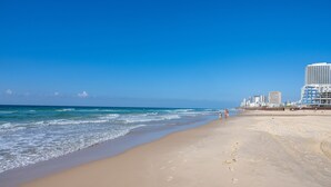 Ein Strand mit blauem Himmel, sandigem Ufer und Gebäuden im Hintergrund.