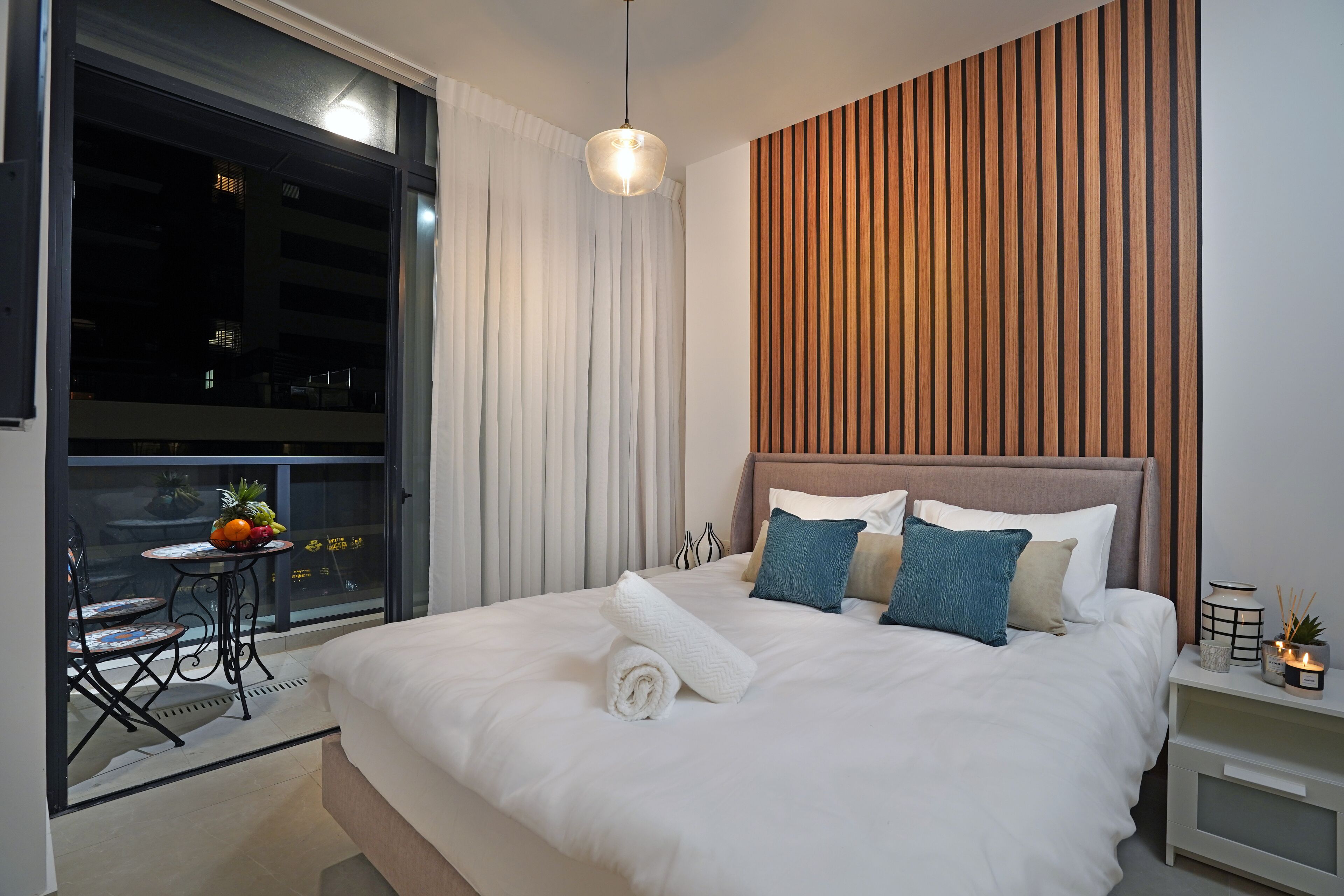 A modern bedroom with a large bed, a wooden panel wall, and a balcony with a table and chairs.