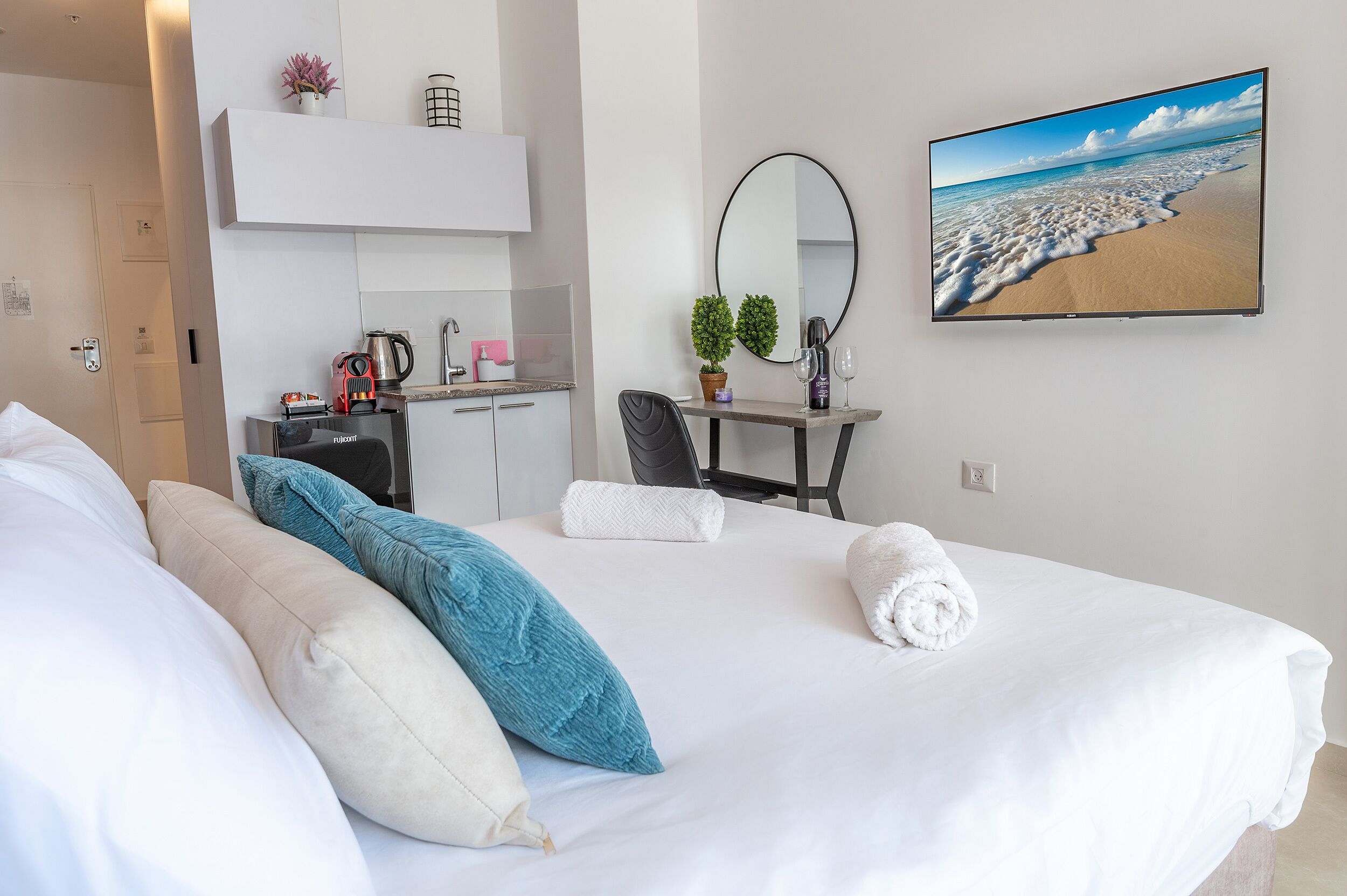 Hotelkamer met een bed, kussens, een bureau met stoel, een koffiezetapparaat, een potplant, een spiegel en een televisie waarop een strand te zien is.