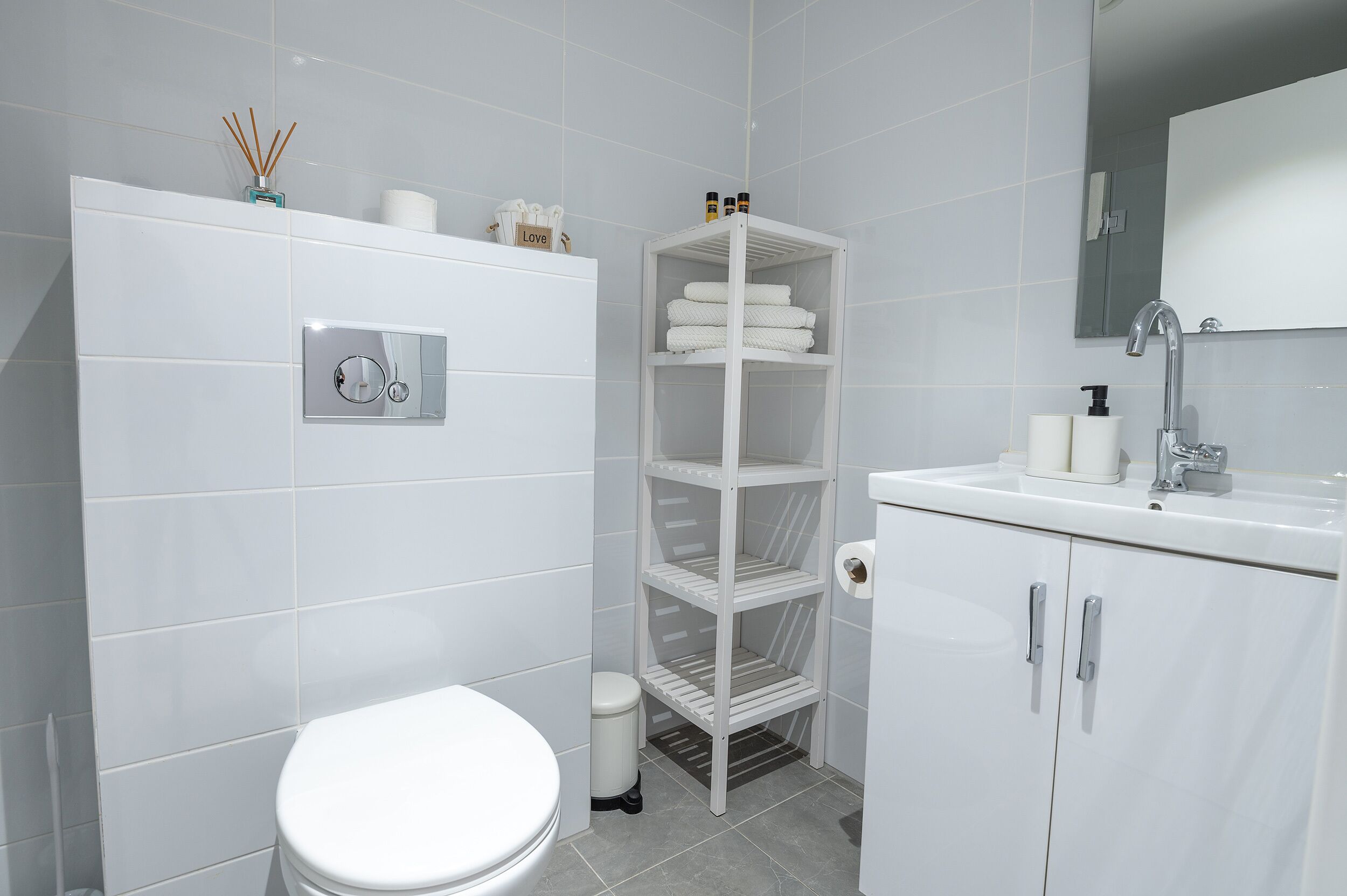 Une salle de bain moderne avec des toilettes, un lavabo blanc avec un miroir et une étagère blanche où sont rangées des serviettes.
