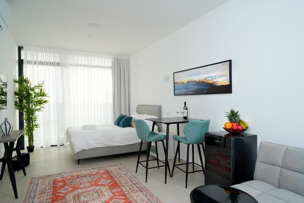A modern hotel room with a bed, a dining area with chairs, a small table with a bottle, a fruit bowl, and a painting on the wall.