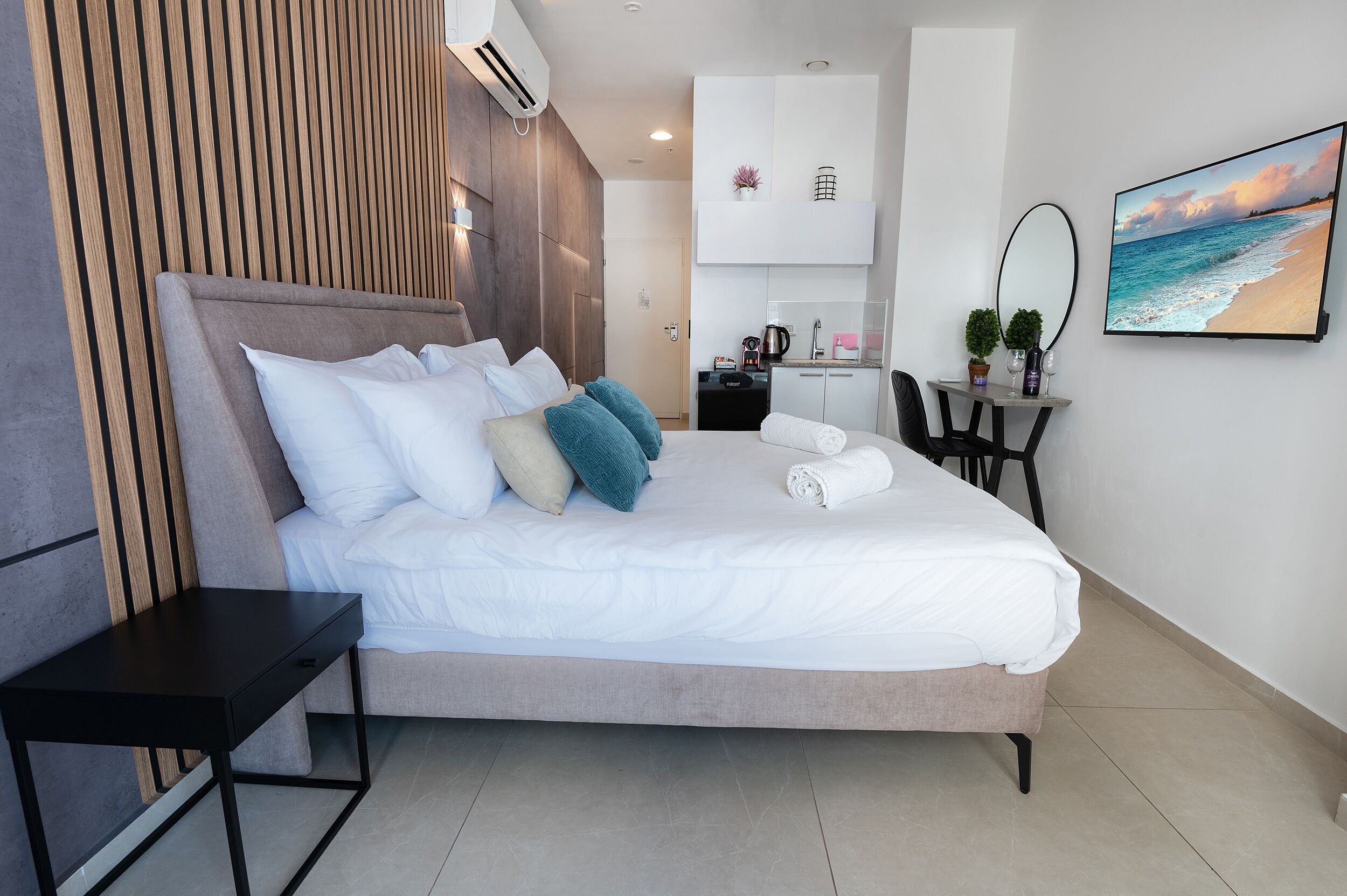 Ein modernes Hotelzimmer mit einem großen Bett, einem kleinen Schreibtisch, einer Sitzecke und einem Fernseher, der eine Strandansicht zeigt.