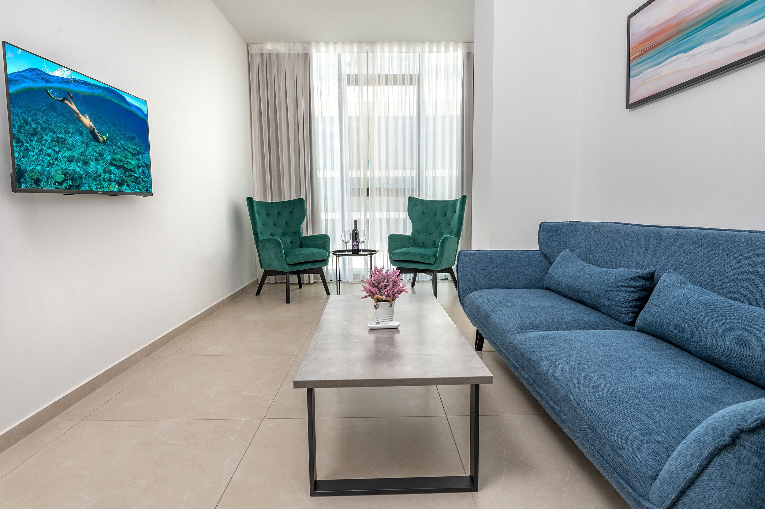 Un salon moderne avec un téléviseur à écran plat, un canapé bleu, deux fauteuils verts et une table basse sur laquelle est posé un vase.