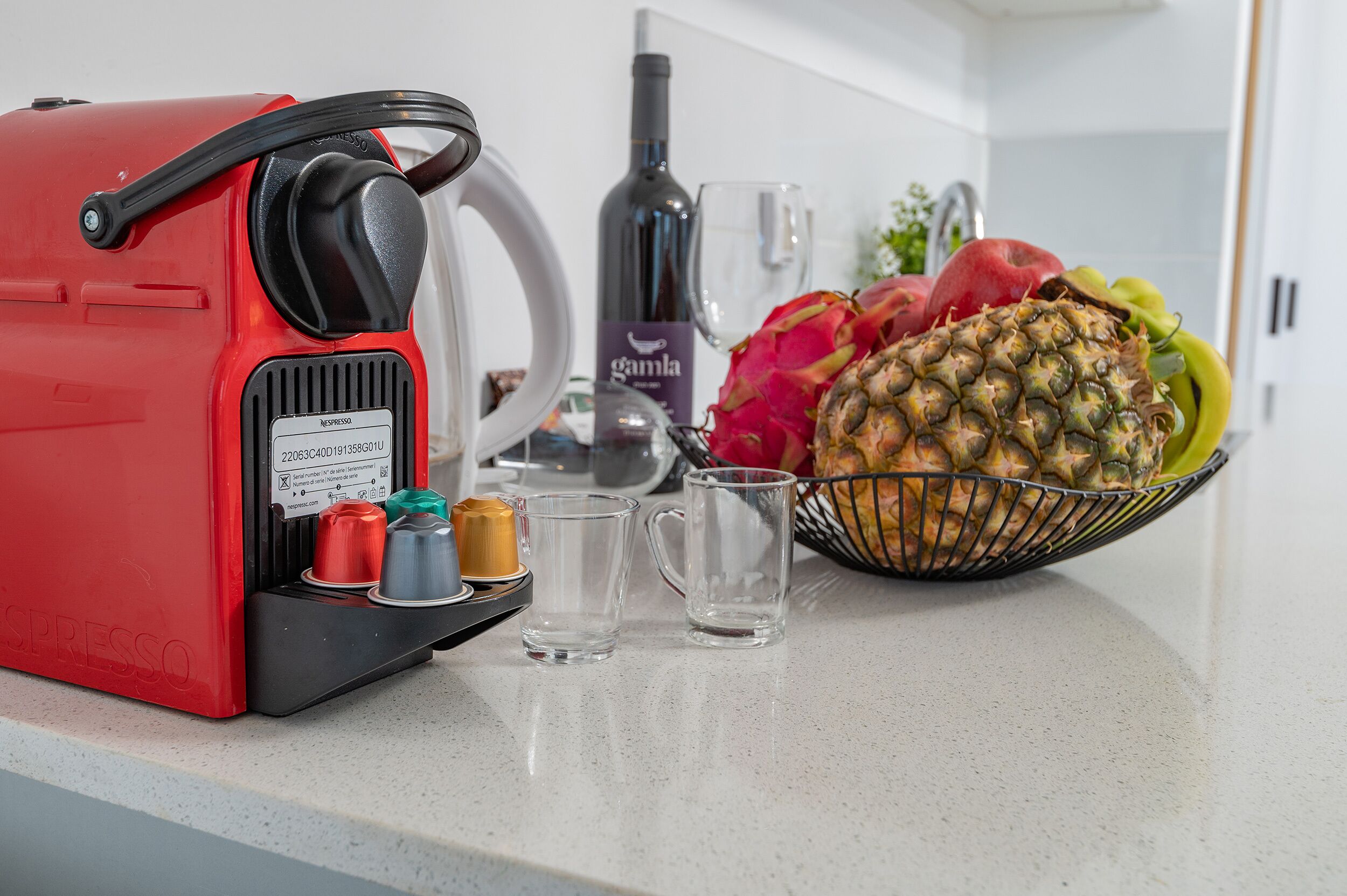 Una máquina de café espresso roja, una botella de vino y un frutero en la encimera de la cocina.