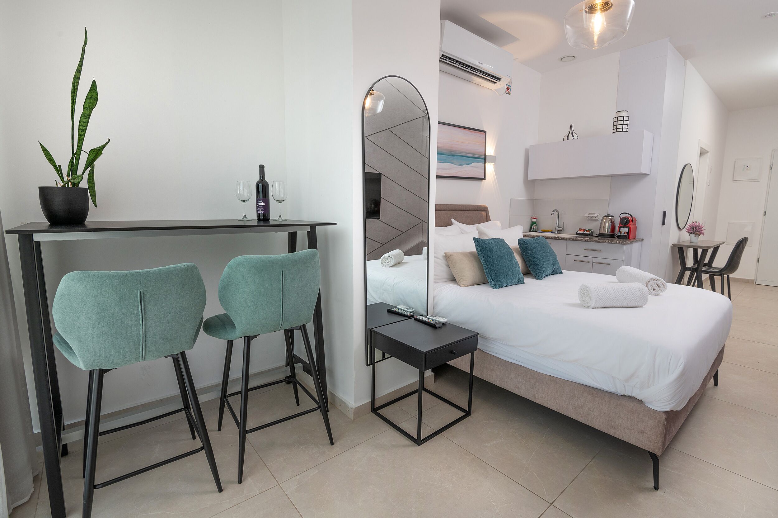 Une chambre d’hôtel moderne équipée d’un lit, d’un bureau avec deux chaises, d’une plante en pot et d’une petite kitchenette.