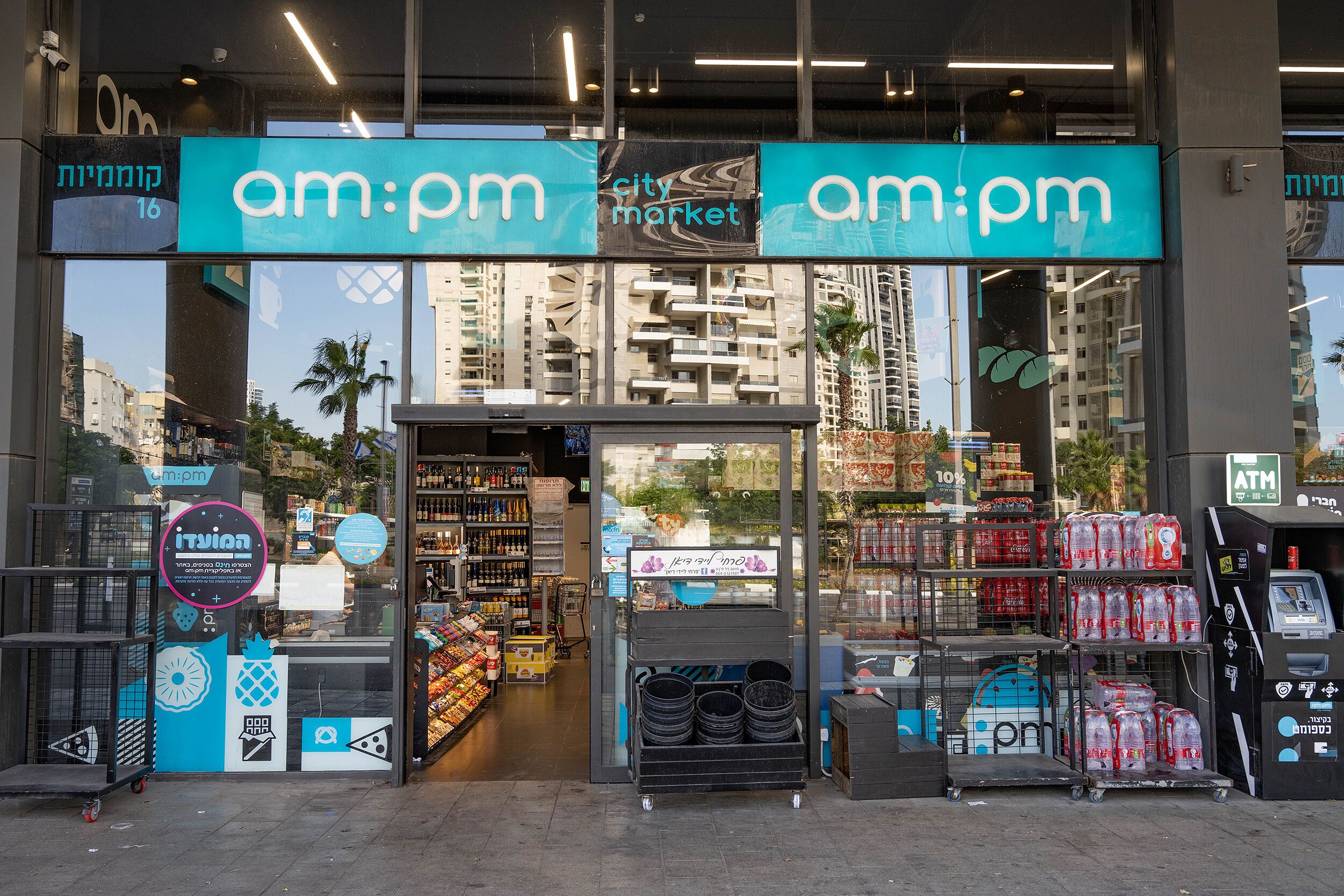 De ingang van een am:pm winkel met een helder zicht op het interieur, waaronder schappen vol producten en een zichtbare geldautomaat.