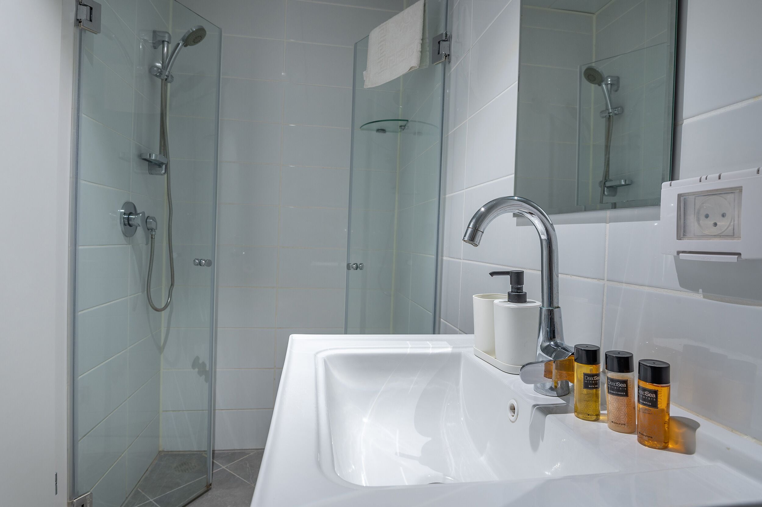 A modern bathroom with a glass shower enclosure, a white sink with soap dispensers, and a mirror above the sink.