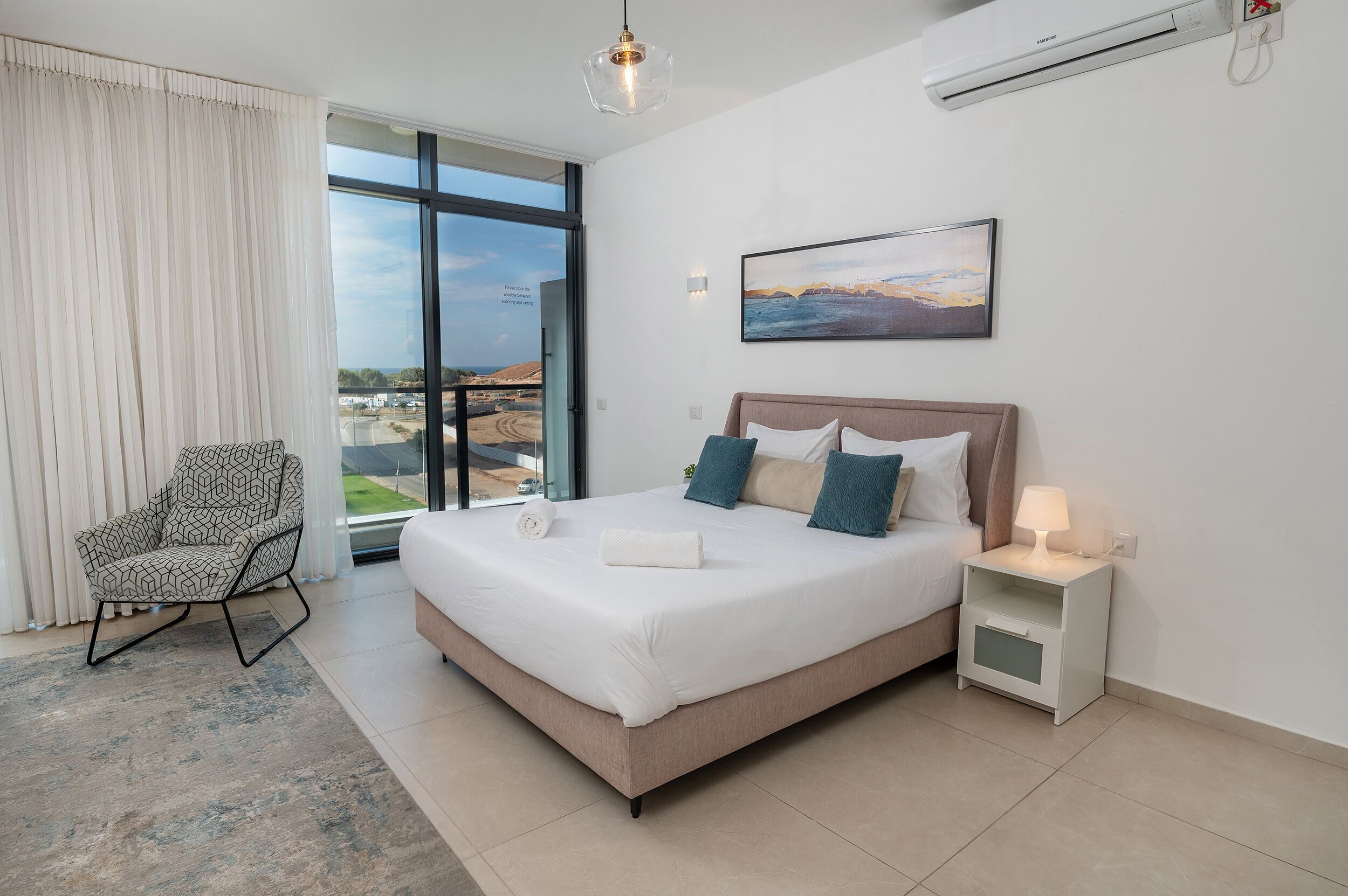 A modern bedroom with a large bed, a chair, a bedside table with a lamp, and a painting on the wall.