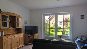A living room with a television, wooden cabinets, a sofa, and a view of a garden through a glass door. - Vacation home in Altfunnixsiel/Carolinensiel (Wittmund)