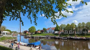 A riverside view with boats docked, a pedestrian path, and houses in the background. - Vacation home in Altfunnixsiel/Carolinensiel (Wittmund)