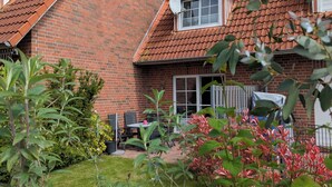 A two-story house with a red-tiled roof and brick walls, a small garden with green and red plants, and a clear blue sky. - Vacation home in Altfunnixsiel/Carolinensiel (Wittmund)