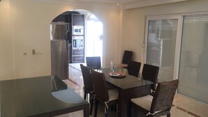 A dining area with a glass table and wicker chairs, a kitchen in the background, and a sliding glass door. - Fully detached Villa offers an unforgettable holiday with your family and friend (Alanya)