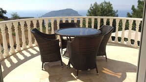 A balcony with a round table and wicker chairs, overlooking a scenic view of the sea and a distant mountain. - Fully detached Villa offers an unforgettable holiday with your family and friend (Alanya)
