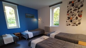 Habitación tipo dormitorio con tres camas individuales, cada una con una almohada y una toalla enrollada. Dos ventanas proporcionan luz natural, y una obra de arte en la pared muestra pájaros y hojas.