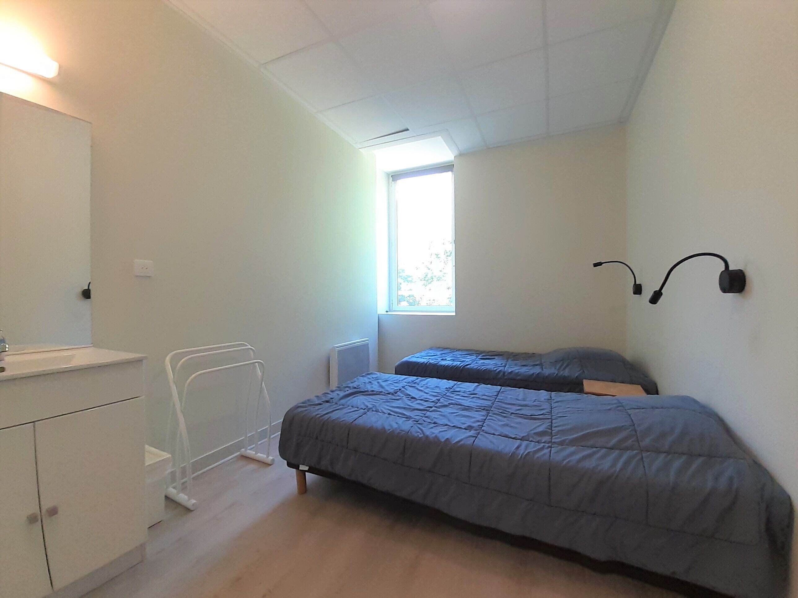 A small, well-lit bedroom with a single bed, a bedside table, a radiator, and a window providing natural light.