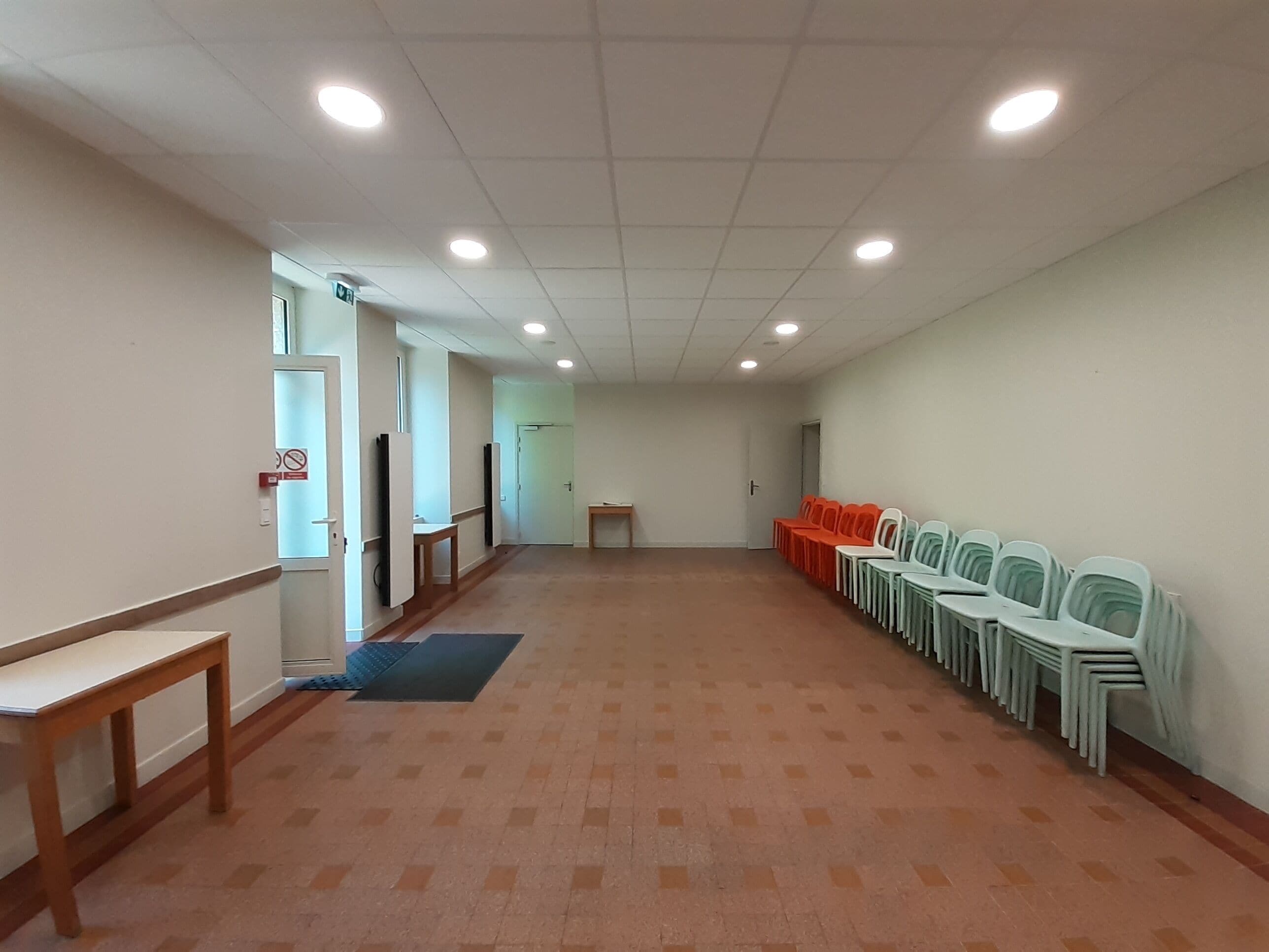 Un hall d’entrée spacieux, avec une longue rangée de chaises d’un côté, un banc en bois et une porte au fond.