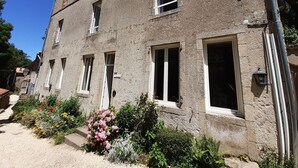 Un edificio de piedra de dos pisos con múltiples ventanas, algunas con contraventanas, y un pequeño jardín con diversas plantas y flores.