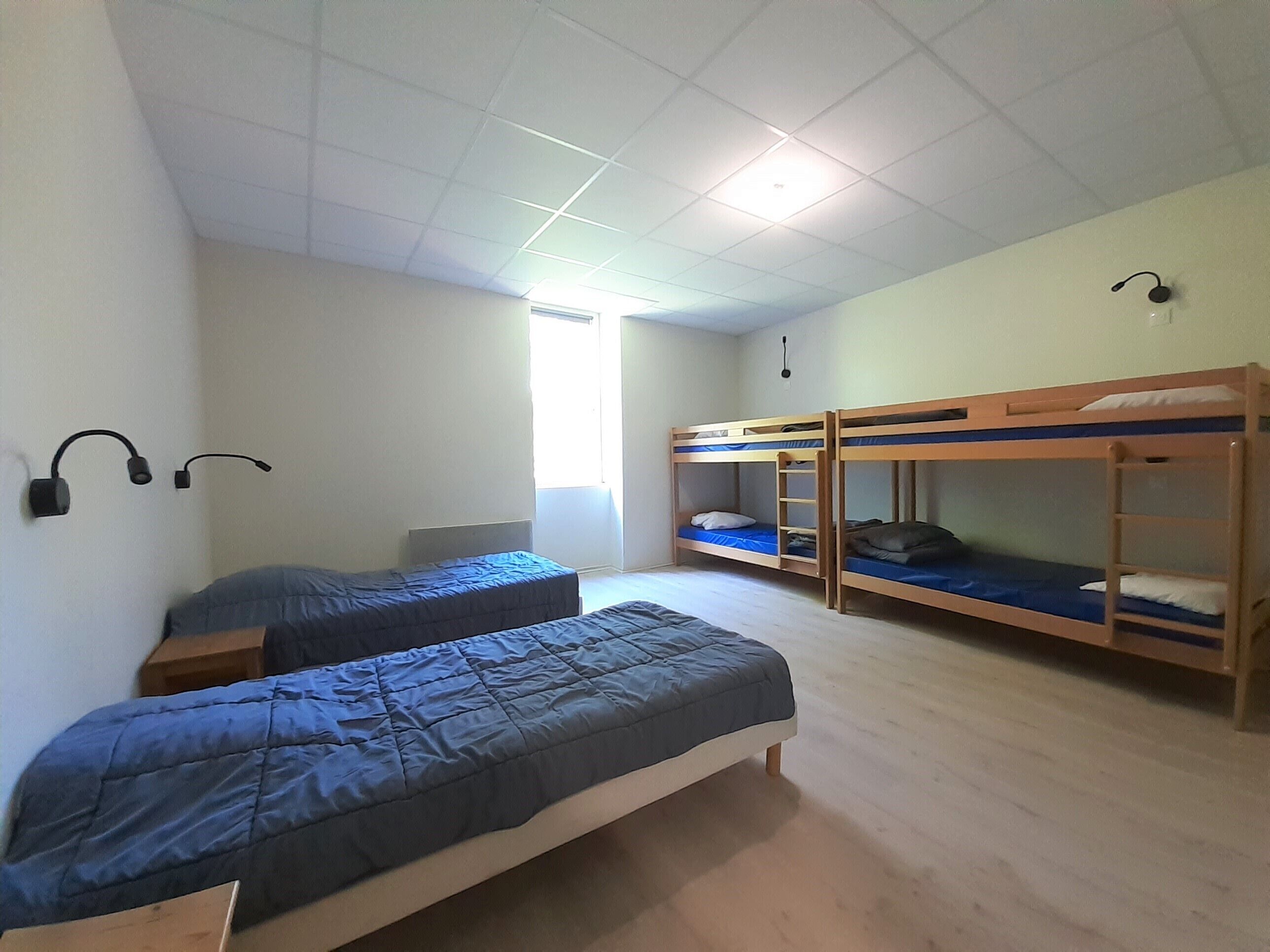 Een slaapzaal met twee eenpersoonsbedden en twee stapelbedden, een plafondlamp en een raam met zonwering.