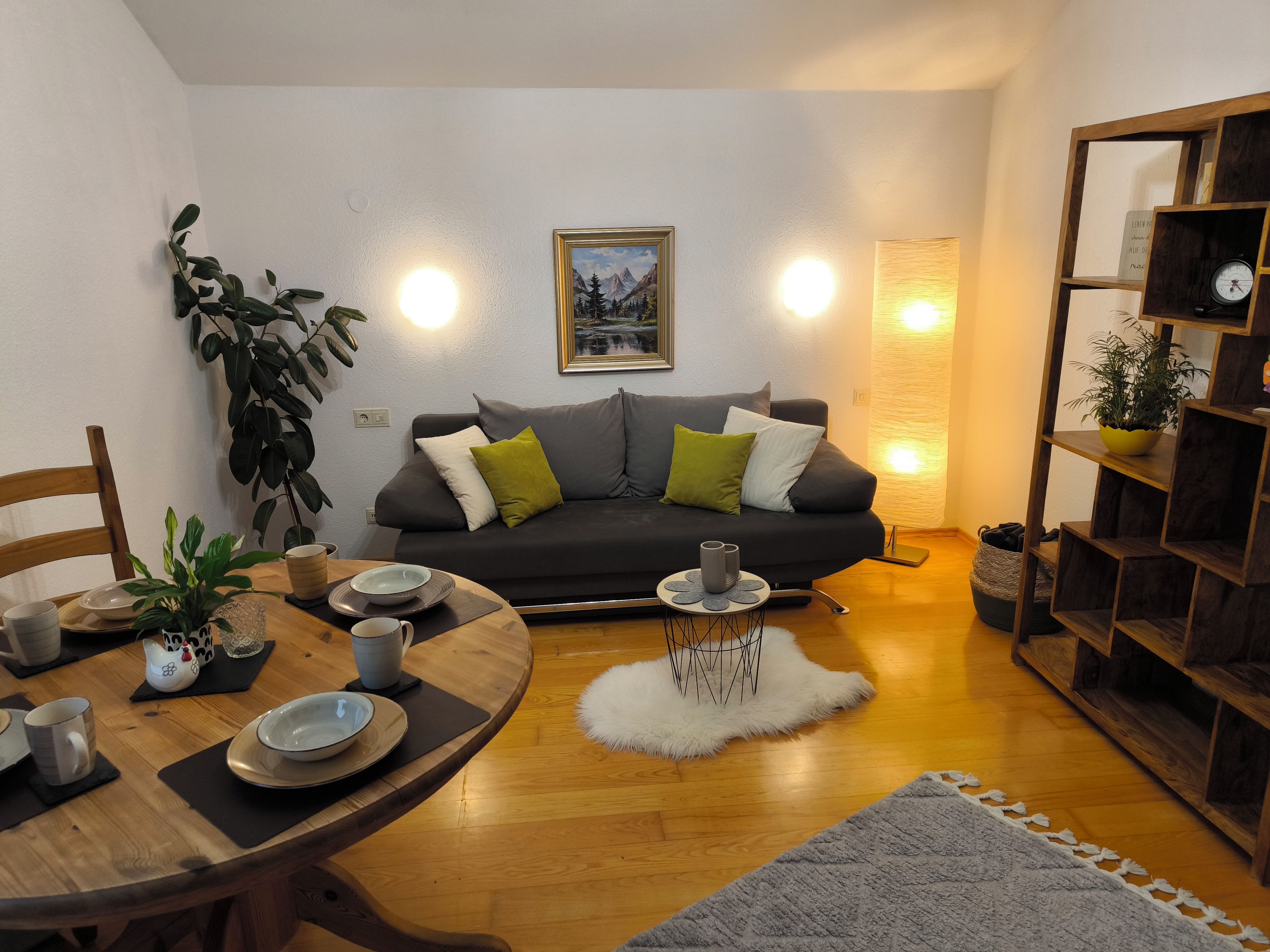 A cozy living room with a dark grey sofa, a wooden dining table, and a bookshelf.
