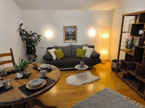 A cozy living room with a dark grey sofa, a wooden dining table, and a bookshelf.