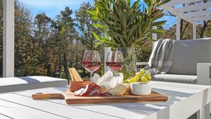 A table with a wooden board holding snacks, two glasses of red wine, a bowl of olives, and a vase with green leaves. - Windermere – stunning and garden setting (Burradoo)
