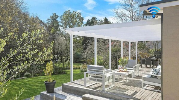 A covered outdoor patio with white furniture, a potted plant, and a view of a garden.
