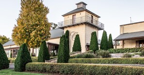 A two-story building with a balcony, surrounded by well-manicured hedges and tall trees.