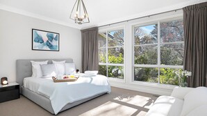 A modern bedroom with a large window, a bed with a white duvet, a bedside table with a lamp, and a framed abstract painting on the wall. - Windermere – stunning and garden setting (Burradoo)