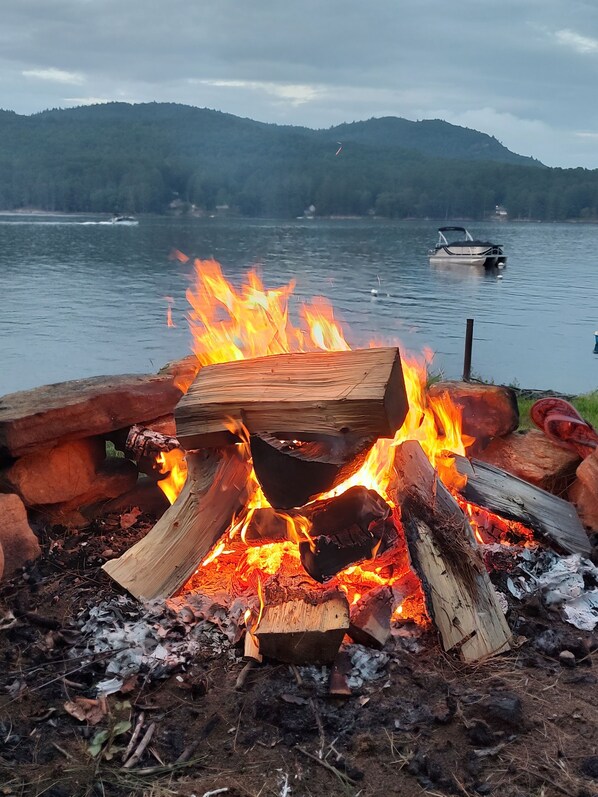 A campfire with logs and a wooden plank, surrounded by natural debris and leaves. - Gorgeous lake view (Hadley)