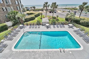 A rectangular swimming pool with lounge chairs and sunbeds, surrounded by a landscaped area with palm trees and a view of the beach. - Cabana One Unit 302 (Myrtle Beach)