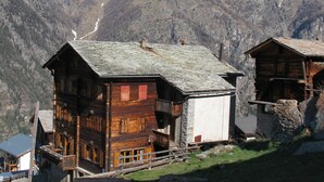 Un chalet de montagne en bois, avec des fondations en pierre, se dressant sur un fond de montagnes enneigées et d’un ciel bleu azur.