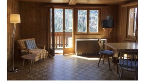 L'intérieur d'un chalet en bois comprend un canapé, une table à manger, un téléviseur fixé au mur et de grandes fenêtres donnant sur la forêt.