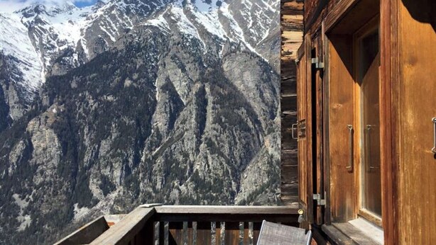 Un balcon en bois, agrémenté d’une table et de chaises, qui offre une vue sur une chaîne de montagnes aux sommets enneigés.