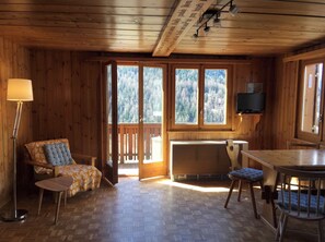 L'intérieur d'un chalet en bois comprend un canapé, une table à manger, un téléviseur fixé au mur et de grandes fenêtres donnant sur la forêt.