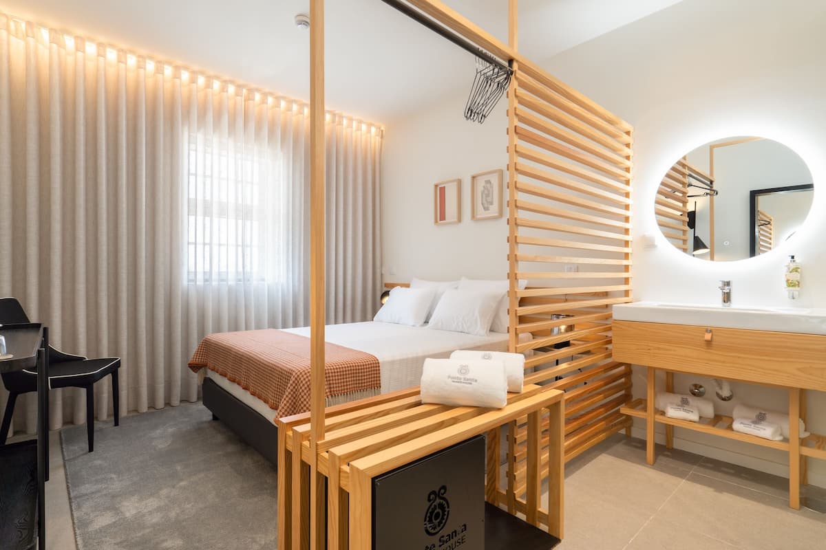 A modern hotel room with a bed, a desk, a chair, a wooden partition, a round mirror, and a bathroom with a sink and towel rack.