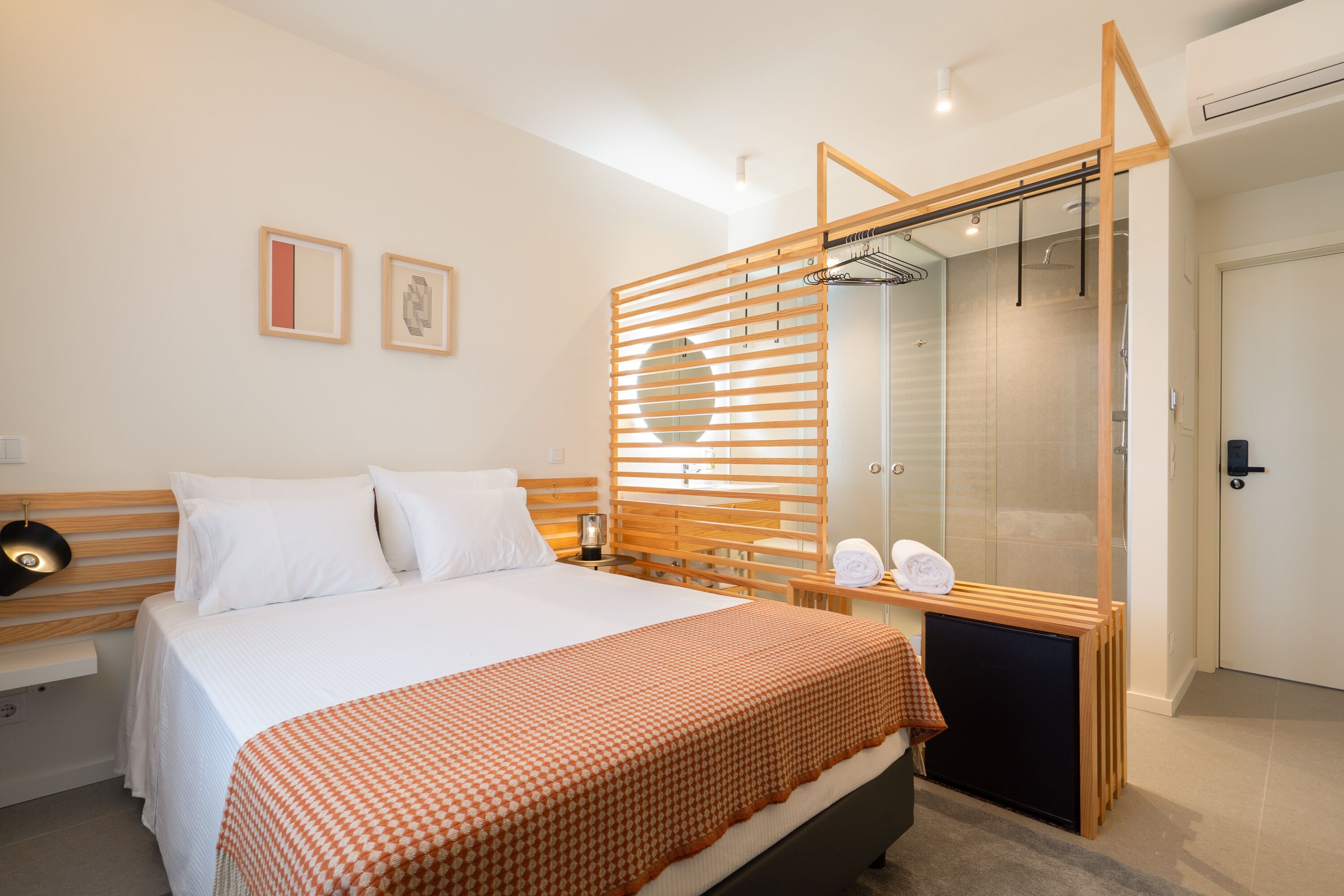 A modern hotel room with a large bed, a wooden headboard, a bedside table with a lamp, a mirror, and a shower area.