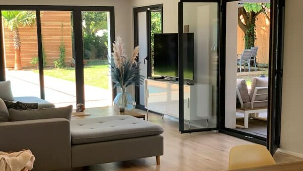 A modern living room with a grey sofa, a wooden dining table, and a TV in the background. - Villa Familiale Centre Bourg sur 2500m² Paysager (Sallebœuf)